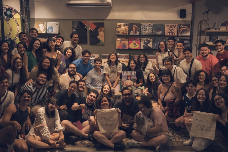 Dezenas de pessoas reunidas e sorrindo para a foto dentro de uma loja de discos, com capas de LP nas paredes e algumas segurando ecobags do Disclubinho.